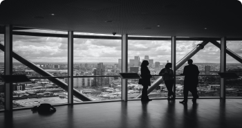three people standing near a panoramic window overlooking skyscrapers